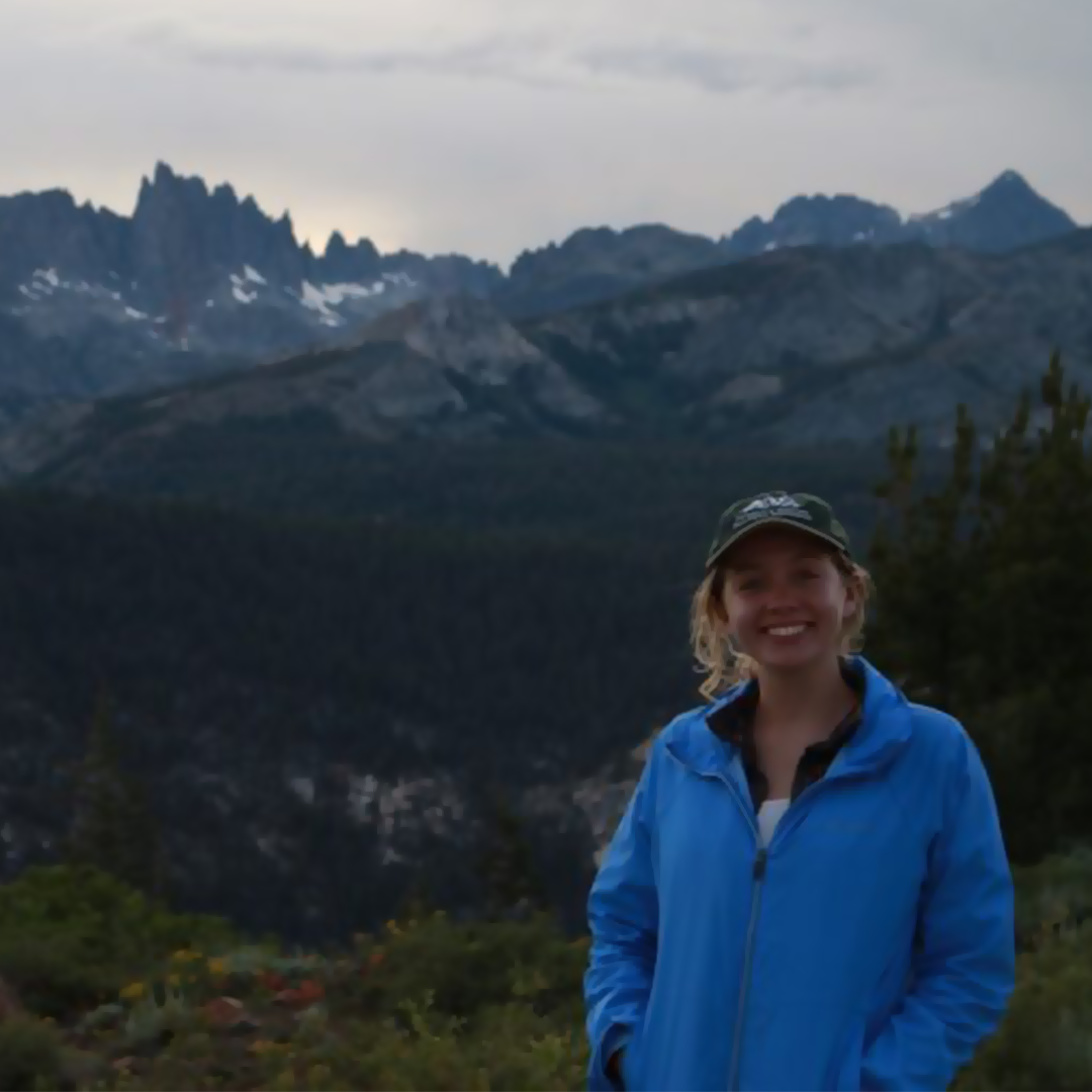 headshot of a white woman with long blond hair and a ball cap with mountains in the background
