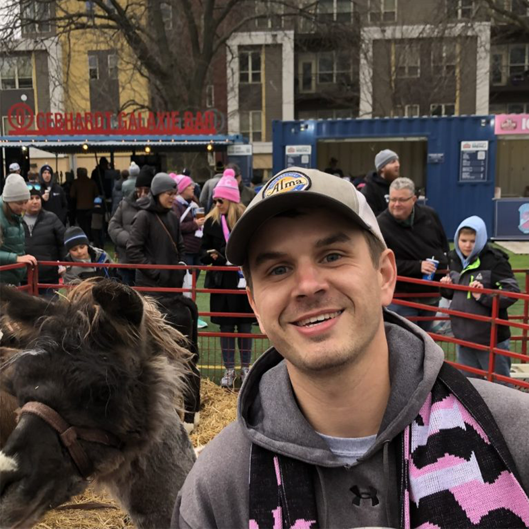 headshot of a white man with a short beard and ball cap with a llama in the background